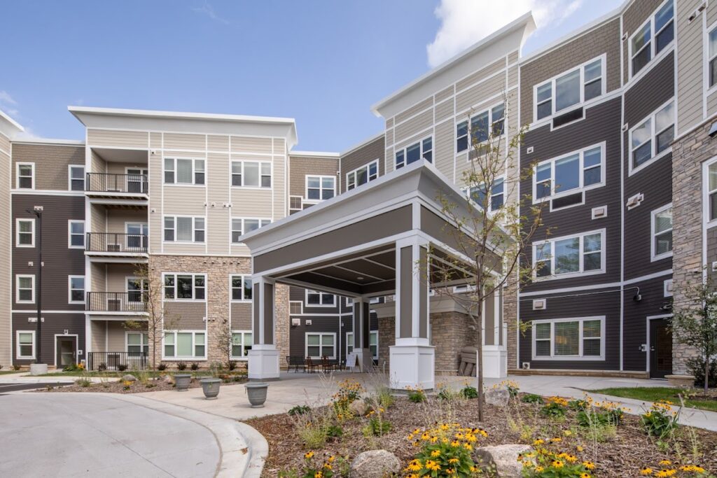 Exterior of a modern apartment building with a covered entrance and landscaped courtyard behind it, including seating under the porte-cochère and colorful flowers.