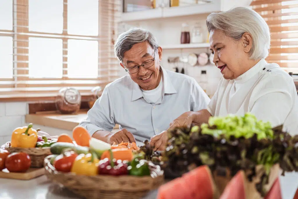 Two smiling elderly adults chopping vegetables together in a bright kitchen with baskets of peppers, tomatoes and greens on the counter.