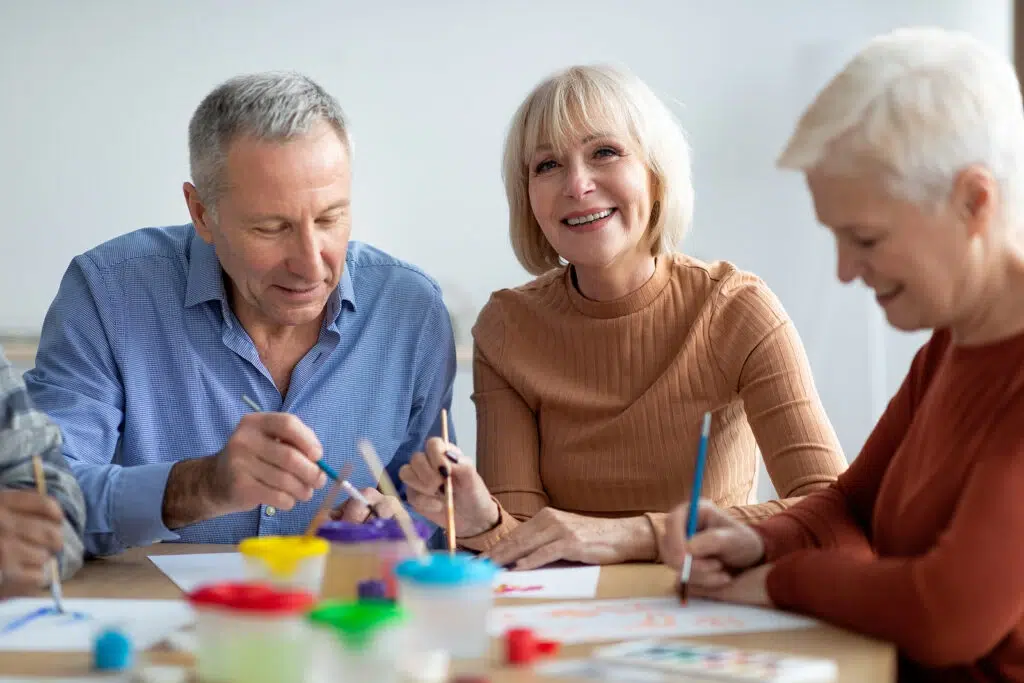 Three seniors painting at a table with bright paints, smiling at the camera.