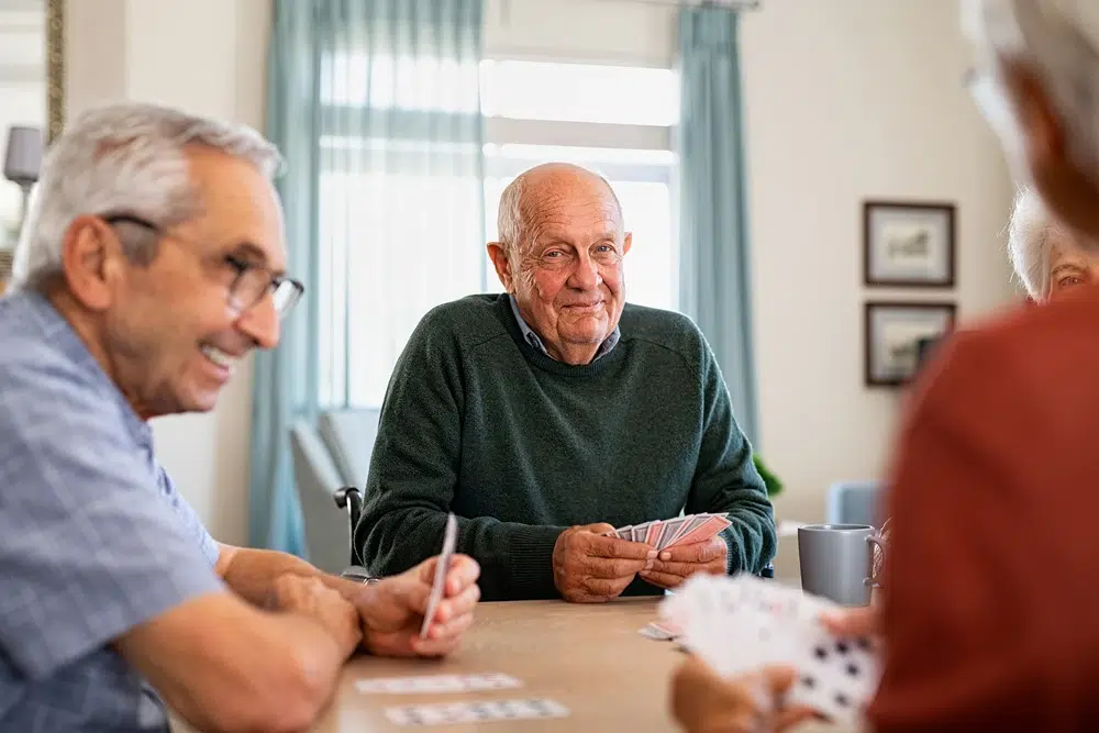 Three elderly adults seated around a table playing cards, smiling as they hold cards in their hands in a bright living room setting.