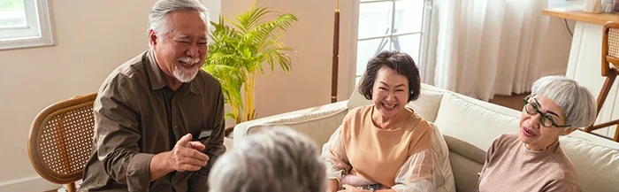 Four seniors seated in a bright living room, laughing and chatting together a friendly conversation around a sofa and chairs.