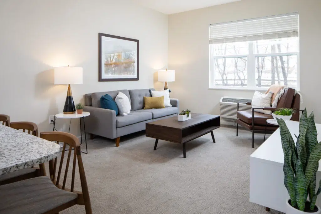 Bright living room with a gray sofa, brown chairs, and a dark wood coffee table under soft lamps and a framed picture on the wall.