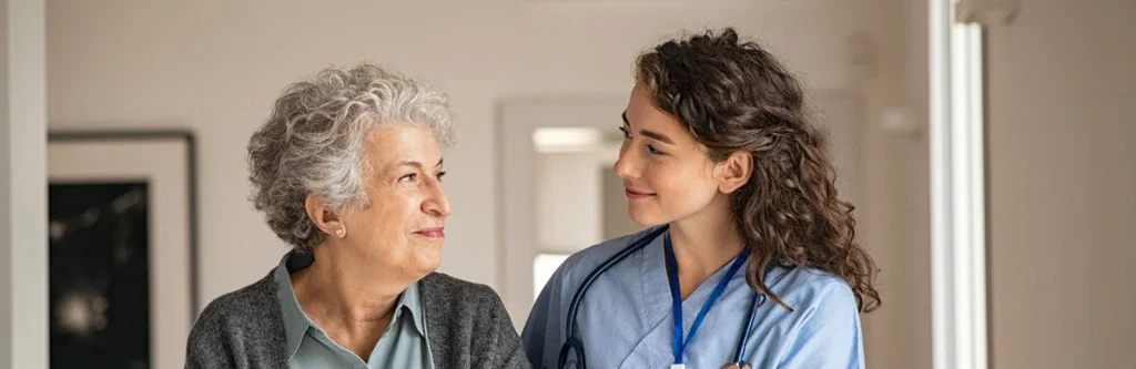 Nurse in blue scrubs helps an elderly woman walk, supporting her arm in a bright hospital hallway.