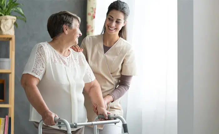 Caregiver helping an elderly woman stand with a walker, both smiling in a bright room.