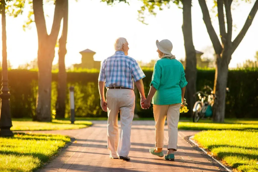 Older couple walks hand in hand along a sunlit park path at sunset, with trees and greenery around them.