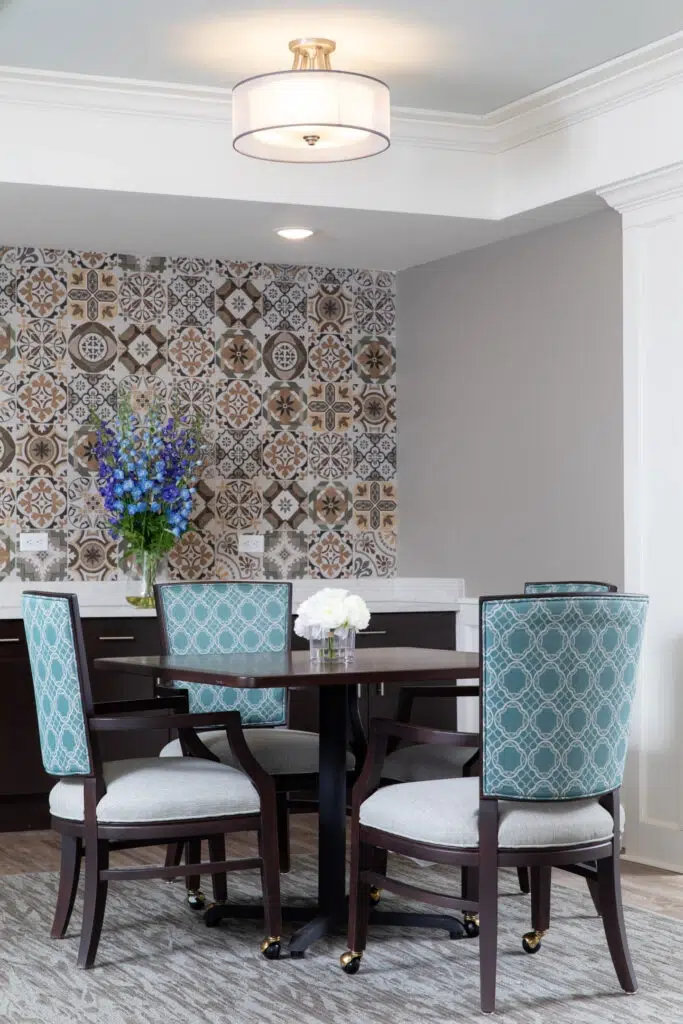 Round dark wooden table with four teal patterned chairs in a dining nook, blue flowers centerpiece and tiled feature wall