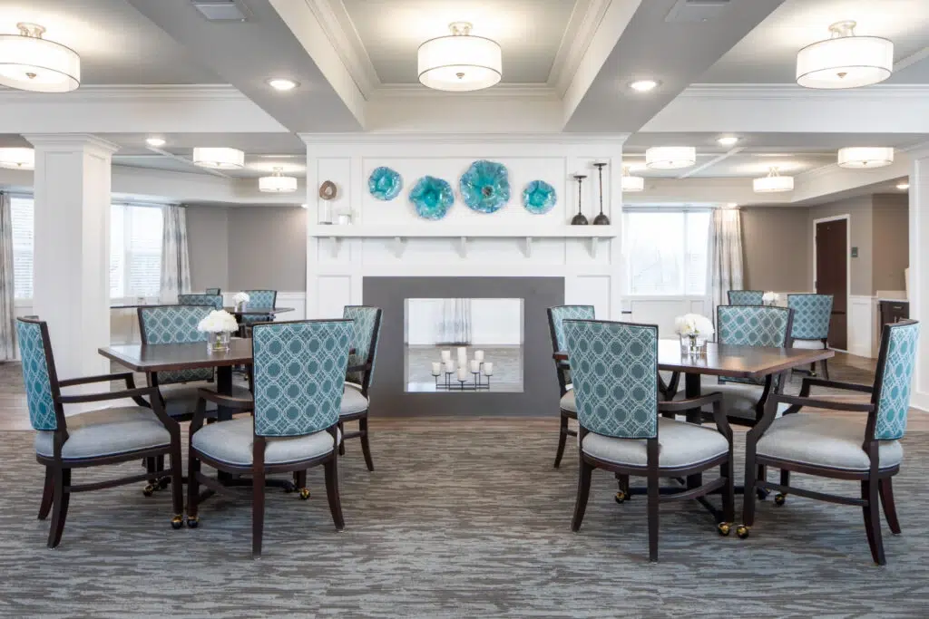 Elegant hotel dining room with round wooden tables and teal patterned chairs around a central fireplace mantel with blue plates above it.