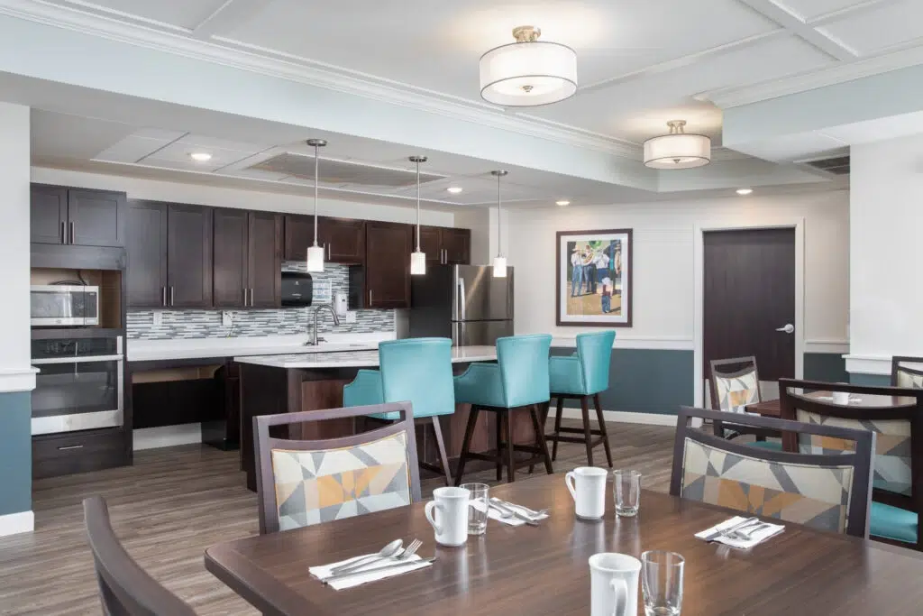 Open‑concept dining area with a dark wood kitchen, stainless appliances, a marble island, and teal upholstered bar stools around the island.