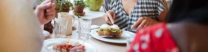 Group of friends sharing a meal at a sunlit table, with a woman in a striped shirt laughing joyfully.