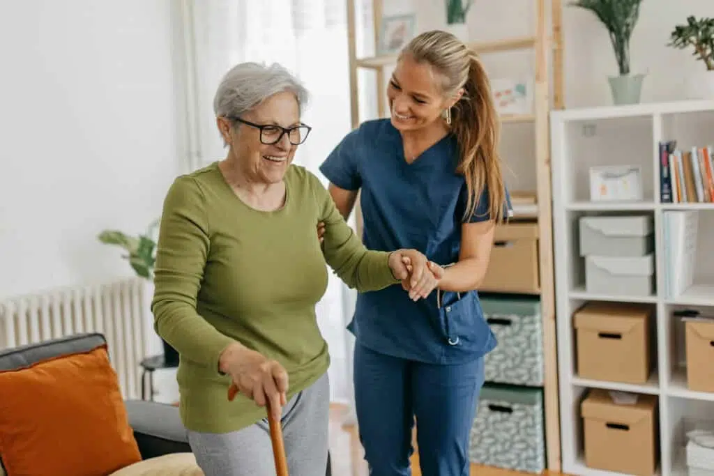 Older woman with a cane receives help from a caregiver in a living room setting.