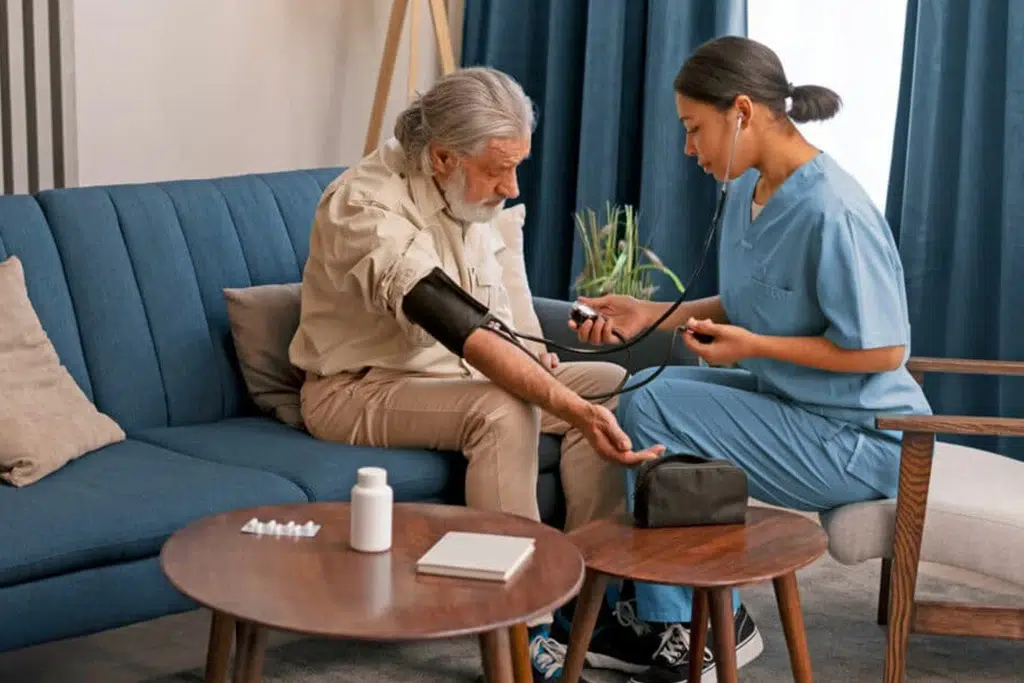 Nurse in blue scrubs checks an elderly man's blood pressure with a cuff and stethoscope at home on a blue sofa.