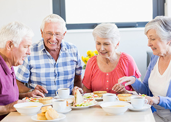 Four senior friends sit around a bright table, sharing a meal and smiling as they converse over bowls, bread, and coffee cups.