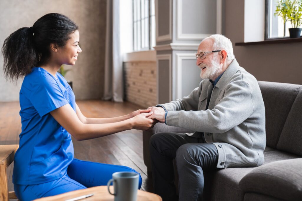 Young female caregiver in blue scrubs holds hands with a smiling elderly man on a couch in a cozy living room.