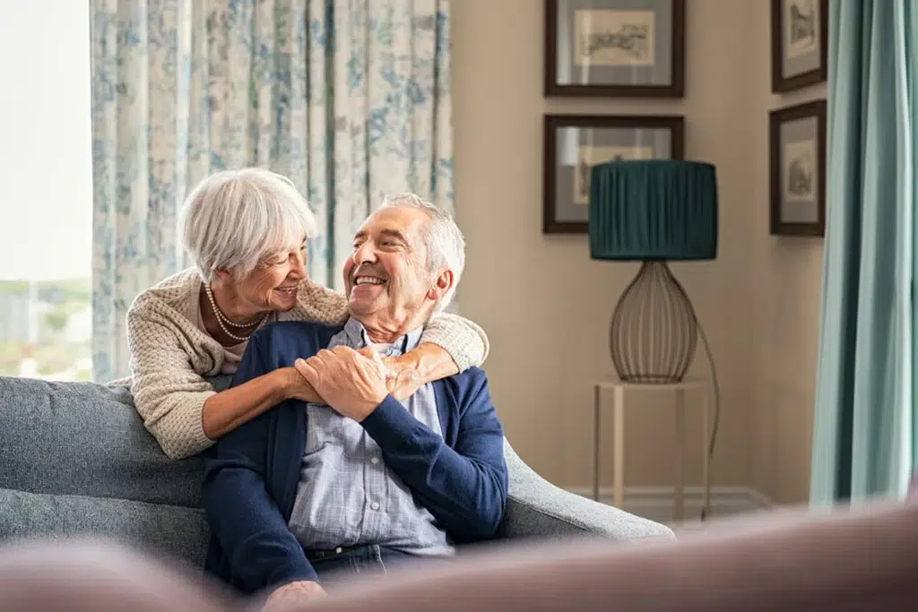 Older couple sitting on a sofa at home, the woman hugging the man from behind as they smile at each other on a cozy living room backdrop.