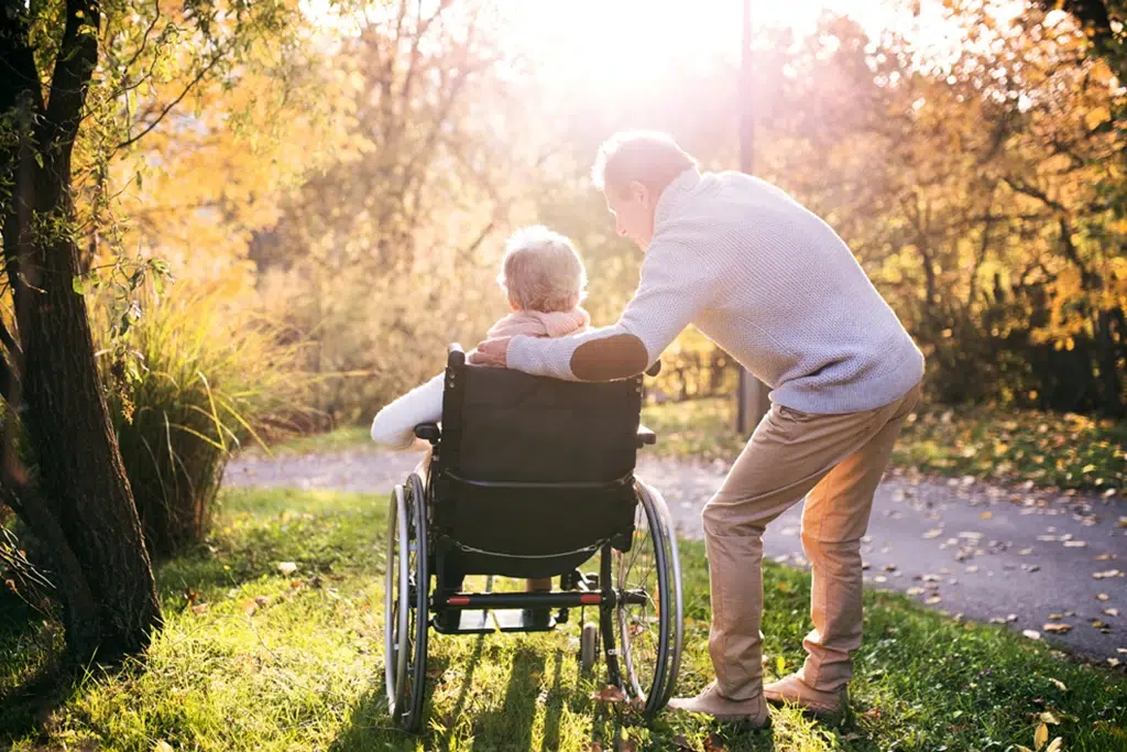 Older person in a wheelchair being assisted by a man along a sunlit autumn park path.