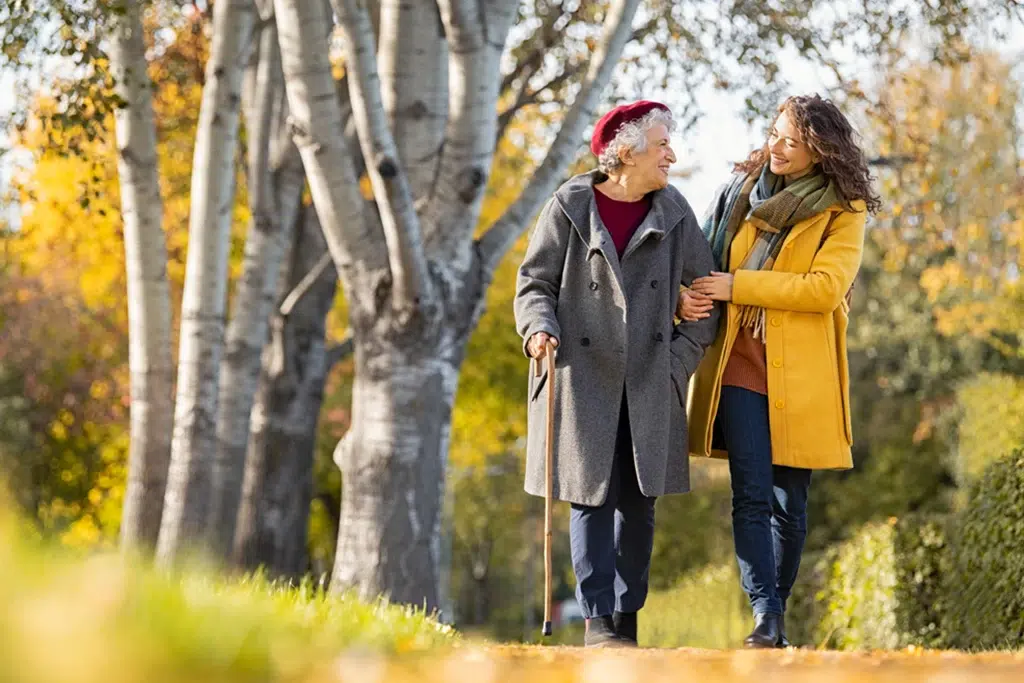 An elderly woman with a cane walking arm-in-arm with a younger woman along a tree-lined autumn path, smiling at each other.