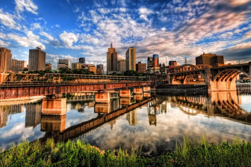City skyline with tall buildings reflected in calm river, bridge on the right and blue sky with scattered clouds.