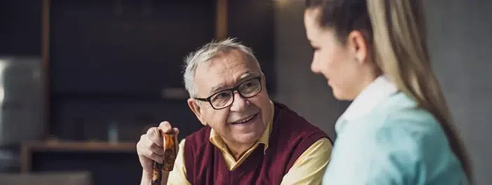 Older man with a cane smiling at a caregiver who is taking notes on a clipboard in a living room setting.