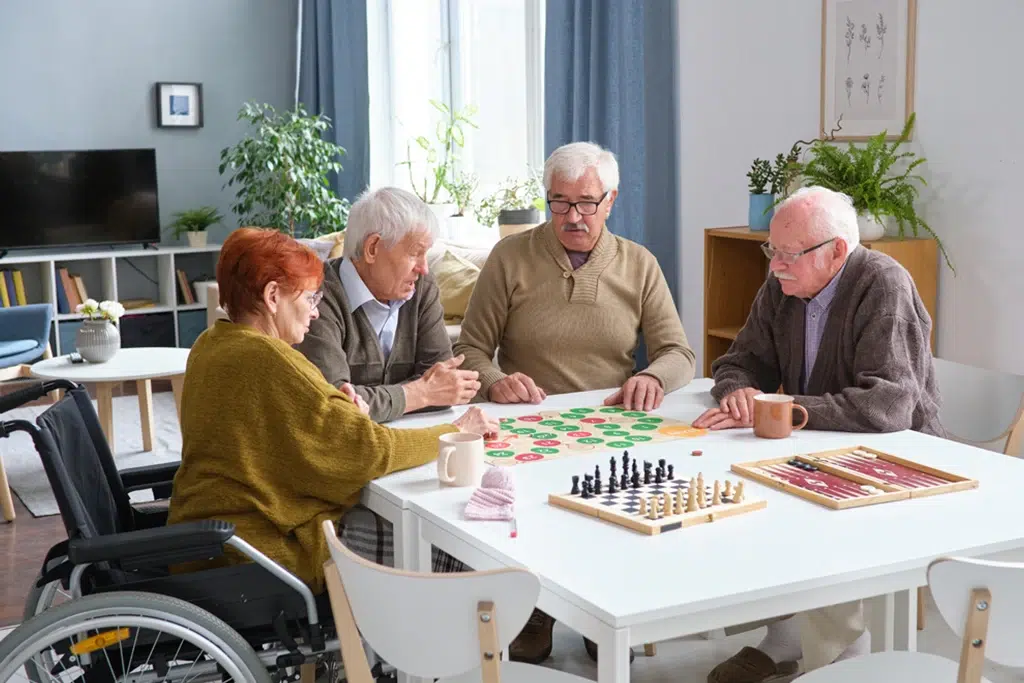 Four elderly people play checkers and chess at a white table in a bright living room; one person uses a wheelchair nearby.
