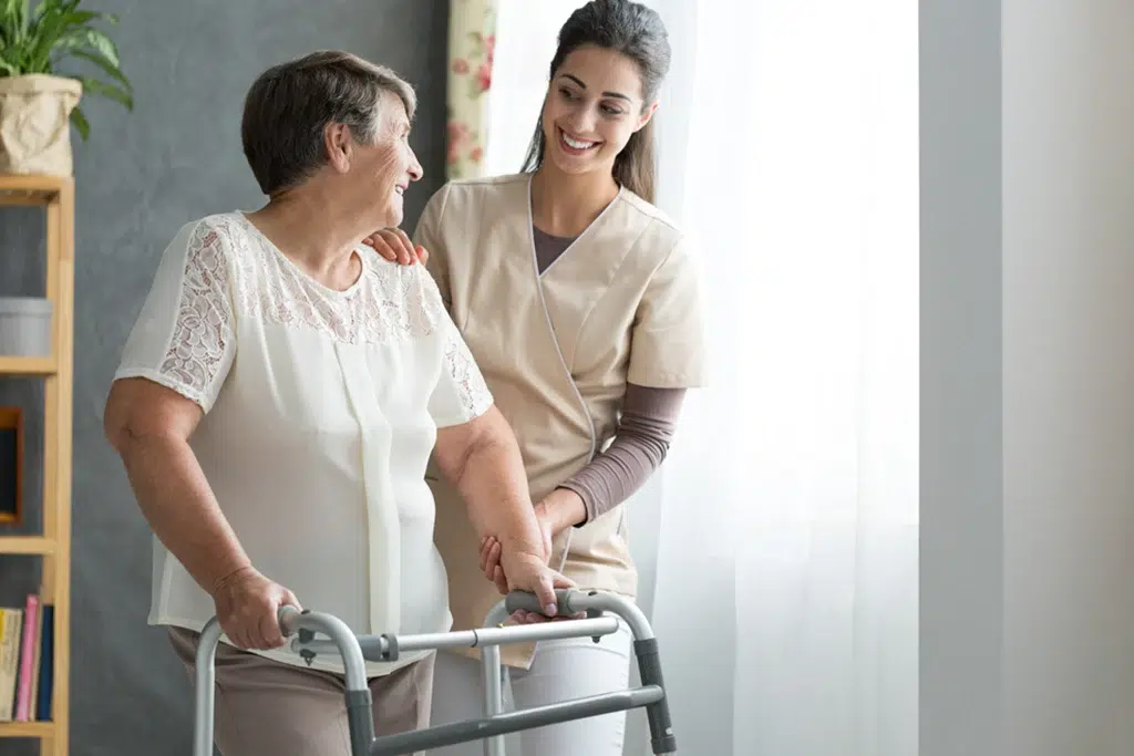Elderly woman using a walker assisted by a smiling caregiver in a beige uniform in a bright room.