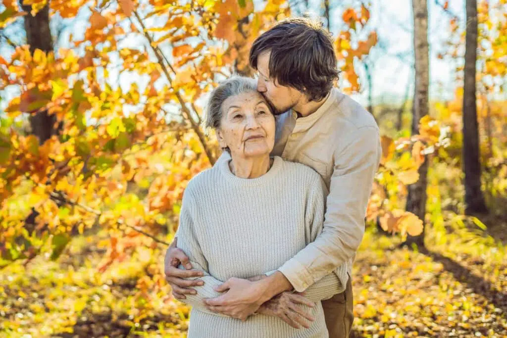 Young man hugging an elderly woman outdoors in a sunlit autumn forest with orange leaves.